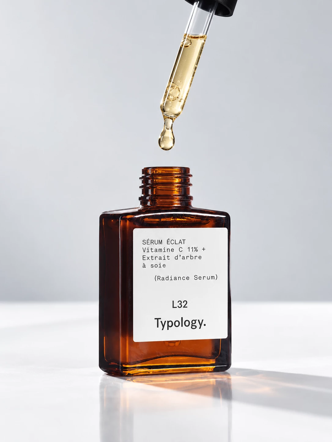 Typology vitamin C serum bottle beneath a glass pipette in soft studio light