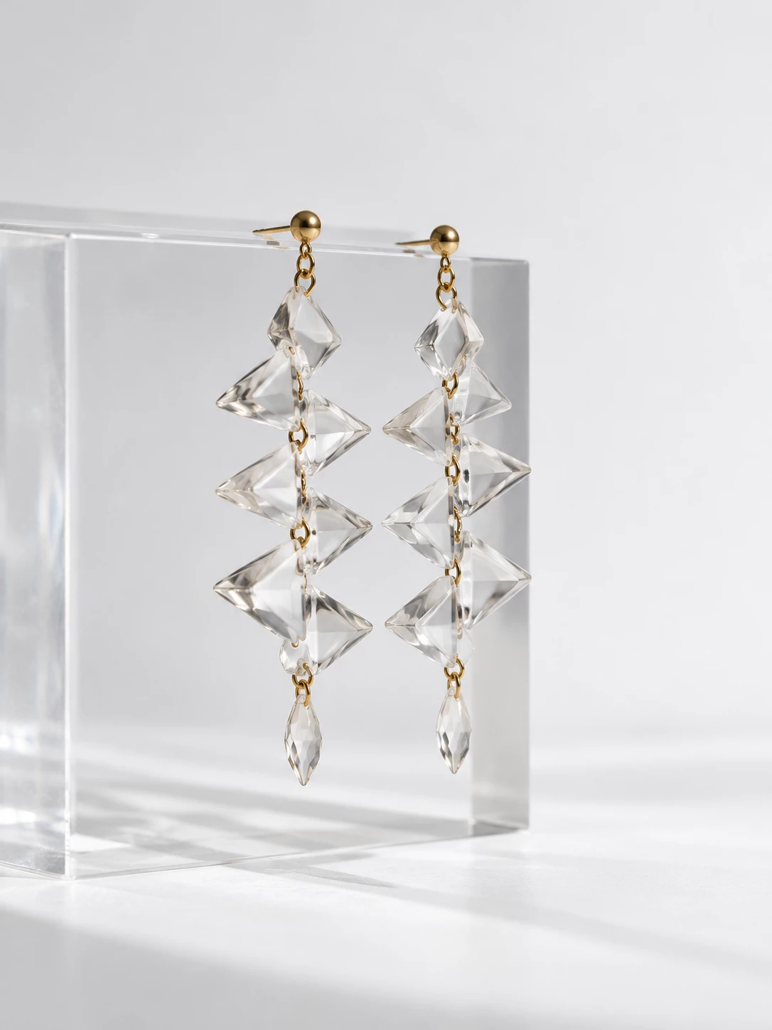 Gold-tone earrings with faceted transparent pendants displayed on a clear glass block