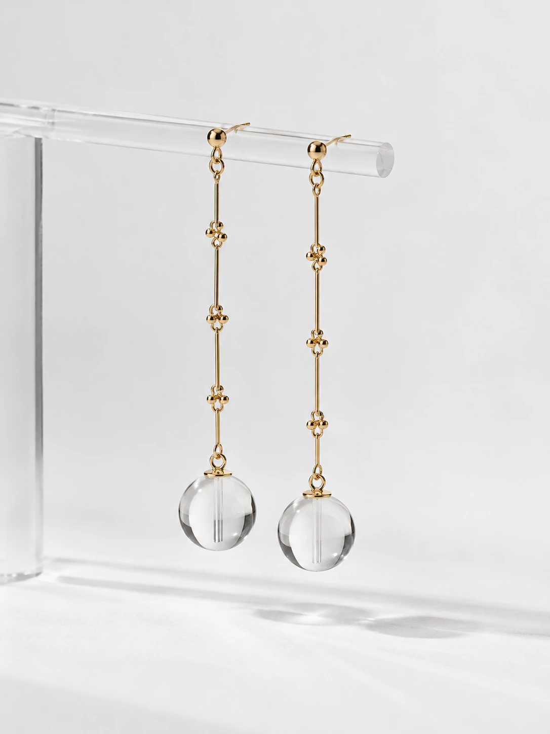 Long gold-tone earrings with clear spherical pendants hanging from a transparent stand