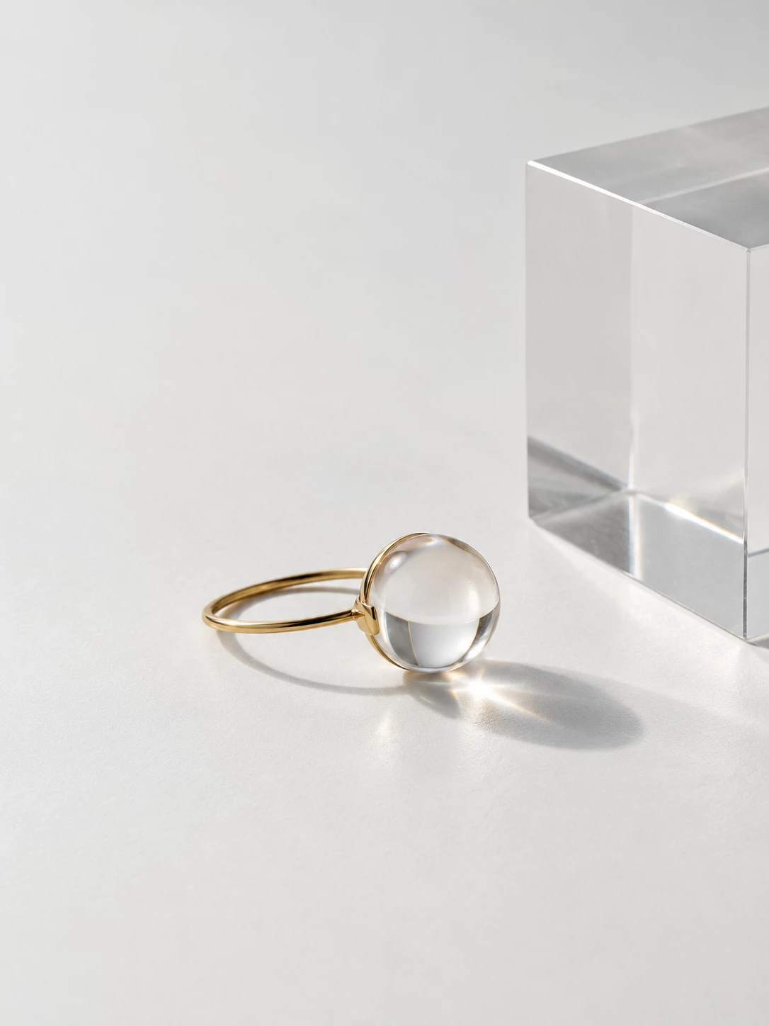 Gold-tone ring with a clear spherical stone beside a glass block in soft sunlight